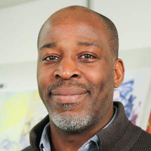 Derrick smiles warmly, wearing a collared shirt. He is a Black man with medium dark skin tone, with closely shaved black hair and a short beard flecked with white.