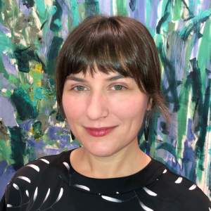Melissa smilles warmly in front of a Joan Mitchell painting. She is a white woman with bobbed brown hair and bangs, and wears a black dress with swooshes of white and a thin gold nose ring..