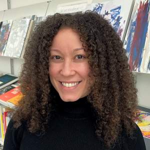 Lauren Galarza smiles in front of a shelf of art books. She has dark shoulder length hair in tight curls, medium-light skin tone, and wears a black turtleneck.