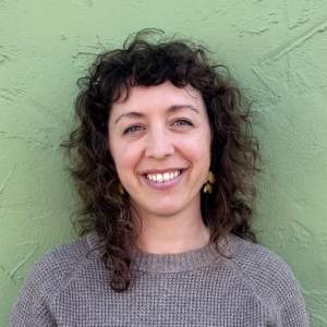 Dani Reuben smiles brightly in front of a green textured wall. She is a white woman with light skin tony, and long curly brown hear, wearing a grey sweater.