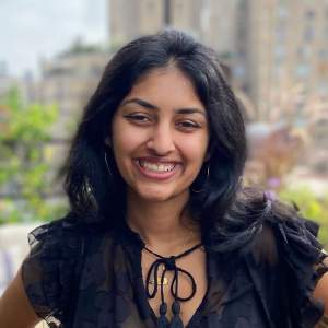 Kaira stands on a rooftop garden in NYC smiling warmly. She has long black hair, a medium skin tone, and wears a ruffled black shirt with a gold necklace.