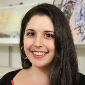 LIlia smiles warmly, wearing hoop earrings and a striped shirt. She is a white woman with light skin tone, and long dark, straight hair.