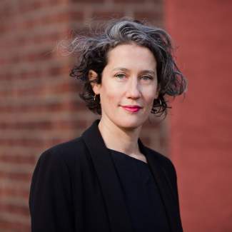 Solana Chehtman, a Latinx woman with curly black and grey hair and a slight smile, wearing a black blazer in front of brick wall