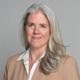 Christa Blatchford, a white woman with long grey-blonde hair and medium-light skin tone, smiles, wearing a tan blazer and collared shirt.
