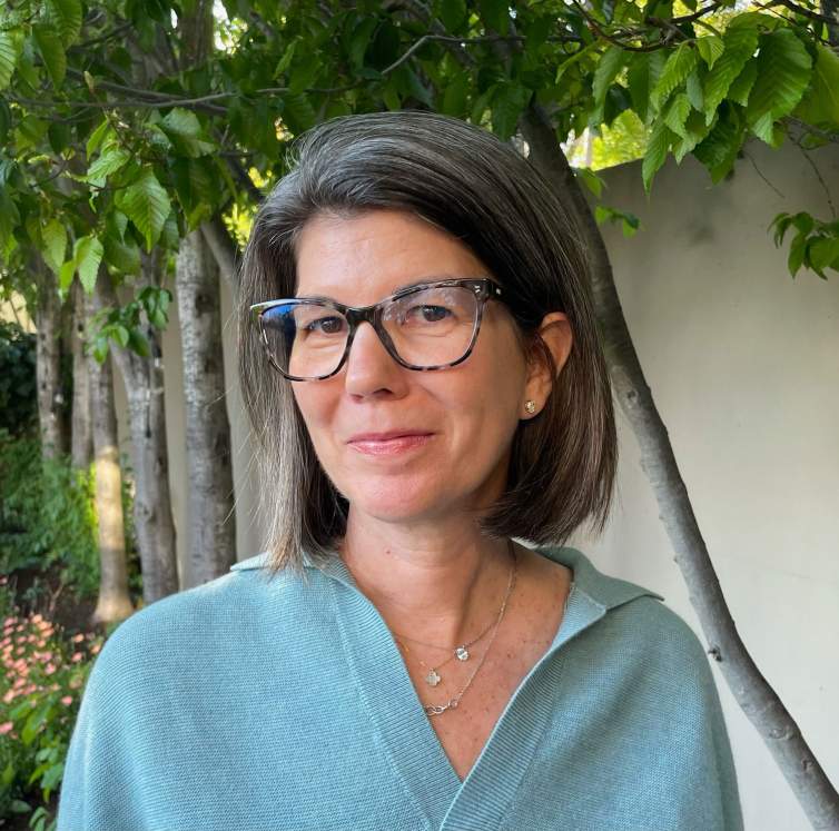 Sarah Roberts smiles in front of greenery. She is a white woman with grey-brown shoulder length hair, wearing glasses and a light blue sweater.