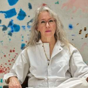 Rebecca Morris sits on a wooden chair wearing a white jumpsuit and sneakers. She is a white woman with light skin tone and long gray hair, with a colorful abstract painting behind her.