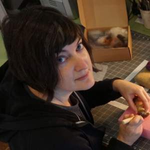ara Engler leans over a work table with a felting needle in her hand, looking over her shoulder to the camera. She is a white woman with light skin tone, and dark chin-length hair, wearing a black hoodie.