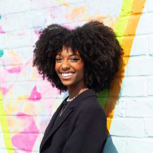Gabrielle Tolliver stands in front of a brightly painted mural. She is a Black woman with a long afro, and she wears a black blazer as she smiles at us from over one shoulder.
