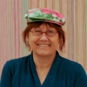 Ayhens, a woman with light skin, smiles in front of a striped wall, wearing a flowered flat cap with red hair peeking out, red wire glasses and a navy shirt.