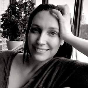 In a black and white photo, Carlson smiles as she leans her head on her hand. She has light skin and long dark hair.