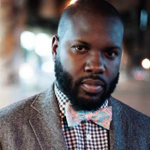 Kasimu, a man with dark skin, a black short beard and short cropped black hair, wears a floral bowtie, checked shirt, and grey jacket.