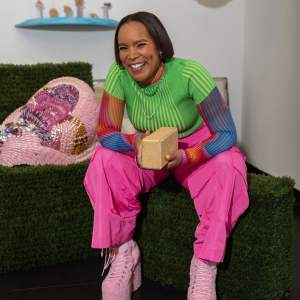Laughing and sitting on an astroturf bench, Council rests elbows on knees, hands holding a tan brick. They have short dark bobbed hair, medium dark skin and wear gold eyeliner, bright yellow nails, bright pink nylon paints, a lime short sleeve knit top over long sleeve multicolor linear patterned sleeves, and light pink boucle platform boots.