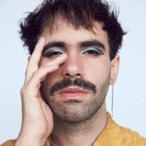 Lucia touches her hand to her face, wearing aqua eyeshadow and a long chain earring. She has short cropped dark hair, a mustache, and light skin.