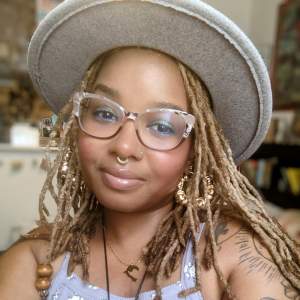 Soraya wears a gray felt hat, gold hoop earrings, septum hoop, and glasses. She has long blonde-brown thin dreadlocks, and medium dark skin.