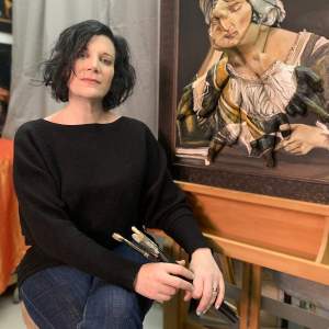 Spector has medium length wavy dark hair and light skin, and sits in front of a figurative painting on a wooden easel, holding several paintbrushes.