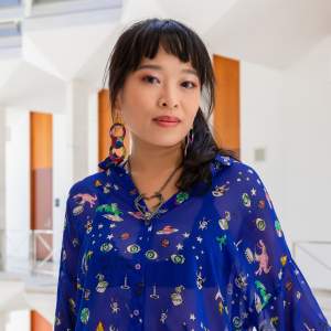 Tsai has long dark hair with bangs, pulled back in a ponytail, and medium skin tone. She wears a royal blue button up shirt with small drawings on it, chunky colorful earrings, and mauve eyeshadow.