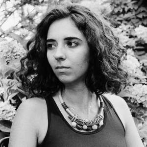 Diane Appaix-Castro looks to the side in a black and white portrait. She is a white and latinx woman with light skin tone and has dark and curly shoulder length hair.
