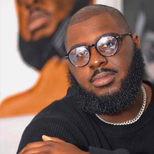 Antony Badon gazes at the camera with hand resting on his arm, wearing black frame glasses, black sweater, nose ring and silver chain. He is a Black man with medium dark skin tone, close cropped black hair, and a full black beard.
