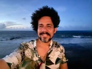 A selfie of Rogelio Báez Vega on a beach at dusk. He is a Puerto Rican man with medium skin tone, curly black hair and a short beard, wearing a tropical shirt.