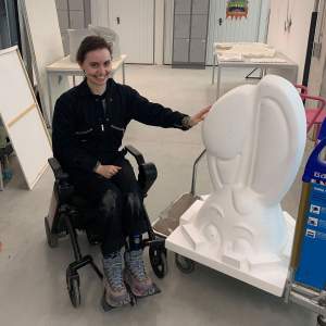 Alcide Breaux smiles with their hand on a large bunny head sculpture. They have short dark hair, light skin tone, are dressed in a dark jumpsuit, and sit in an assistive power wheelchair.