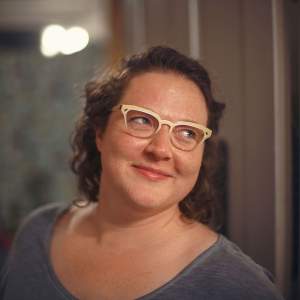 Ellen Bull smiles slightly, and looks up to the right. She is a white woman with light skin tone with freckles, light brown curly hair pulled back, and wears pearl cat eye glasses.
