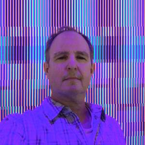 James Flynn stands in front of a patterned painting bathed in pink light. He is a white man with light skin tone, short brown hair, wearing a plaid button up shirt.