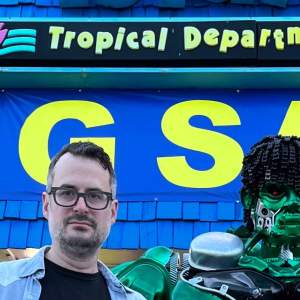 Thomas Friel stands in front of a brightly colored storefront next to a green robot. Friel wears black frame glasses and has light skin tone, black and gray hair and a short beard.