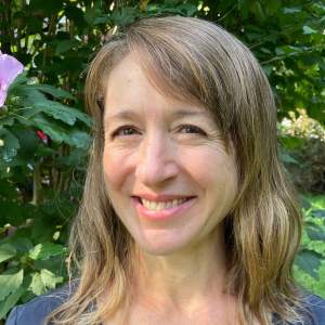 Emily Holt smiles in front of greenery. She is a white woman with light skin tone, and shoulder-length dark blonde wavy hair.