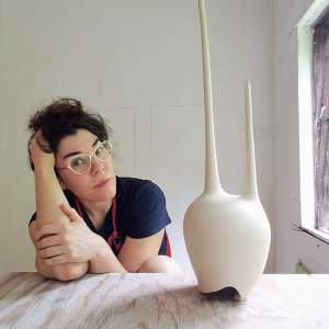 Sarah House leans her head on her hand on a table, next to a pointed abstract ceramic sculpture. She is a white woman with light skin tone, dark short wavy hair, and wears metal frame glasses.
