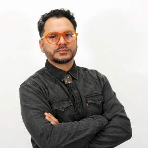 Salvador Jiménez-Flores stands with his arms crossed, wearing orange frame glasses, black denim shirt and bolo tie. A Latino man with medium skin tone, he has short dark hair and beard flecked with gray.