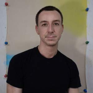 Ryan Leitner sits in front of a pinned-up canvas, wearing a black t-shirt. He is a white man with light skin tone, and short cropped black hair.