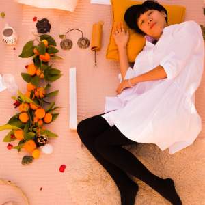 Love Nyguen lays on a sheepskin rug with her head on a marigold pillow in a white dress and black stockings. She is a Vietnamese-American woman with dark short hair, and medium light skin tone, and to her left are oranges with leaves and other objects.