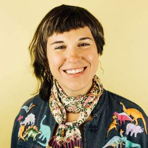 Jane Tardo smiles brightly, wearing a patterned scarf, and a denim shirt with dinosaur appliqués on the shoulders. They are a white person with light skin tone, and have brown straight hair in an asymmetrical cut and bangs.