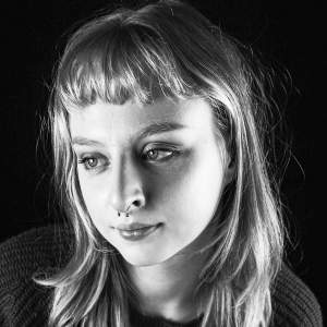 Katya Vaz gazes down in a black and white photo. She is a Russian-American white woman with light skin tone, and long blond hair with short bangs, and a septum piercing.