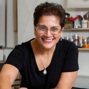 Anila Quayyum Agha smiles brightly at a work table in her art studio. She has short dark hair and glasses, wears a black shirt and necklace, and has a medium light skin tone.