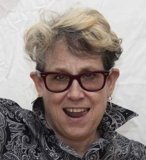 Jerri Allyn smiles impishly. A white woman with medium light skin tone, shw  has short, wavy blonde hair and wears chunky tinted glasses,  a patterned shirt and hoop earrings,