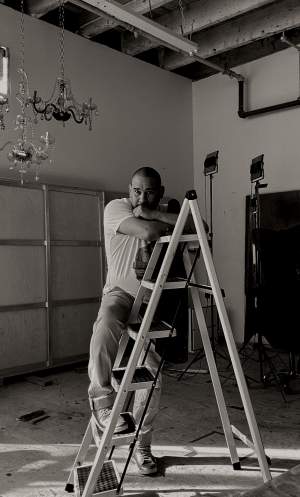 David Antonio Cruz leans on a ladder in a room with exposed ceiling beams, chandeliers, and lighting equipment. He is a Latino man with closely shaved black hair.