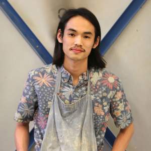 Christian Việt Đinh stands in front of a blue X, wearing a faded clay-splattered blue apron and floral shirt. He is a Vietnamese-American man with dark shoulder length hair, and a mustache.