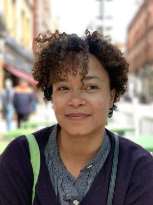 Addoley Dzegede is a Ghanaian-American person with short curly hair and a medium light skin tone, and wears a striped shirt and a cardigan, sitting outdoors, gazing to the side. Background shows a blurred city street with buildings and people.