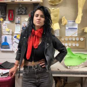 Bethany Grabert stands in a studio space with body castings on the wall. She has dark wavy hair and medium skin tone, and stands with her hand on hip, wearing a black leather jacket, dark jeans and a red scarf, with tattoos visible on her midriff and arm.