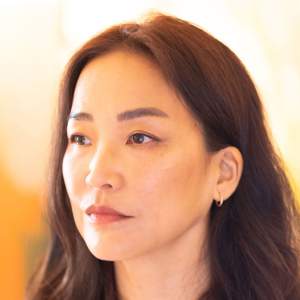 Seonna Hong looks to the side in a light filled room. She has shoulder length brown hair, light skin tone, and wears small gold hoop earrings.