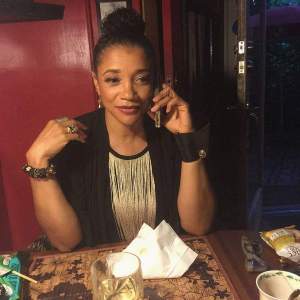 J.Renee, a Black woman with medium skin tone, sits at a table, on the phone, with snacks and a drink nearby. She has short hair and is wearing a black and white outfit.