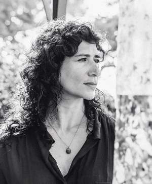 Ivelisse Jiménez gazes to the right in a black and white photo. A Puerto Rican woman, she has long dark curly hair, and wears a dark collared shirt and pendant necklace.