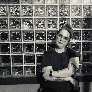 E Marshall stands in front of a glass block wall with tattooed arms crossed. He is a white person with light tone skin and wears glasses, a knit hat, and a black tshirt.