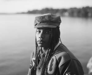 In a black and white photo, a Black woman wearing a newsboy cap and braided hair faces the camera, with a calm body of water in the background.
