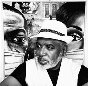 In a high-contrast black-and white image, a Black man with a white beard, wearing a white hat and vest, poses in front of a painting featuring two masked faces.