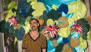 A white man with light skin tone, short brown hair, a mustache, and a striped shirt, stands smiling in front of an art installation of fabric plant life.