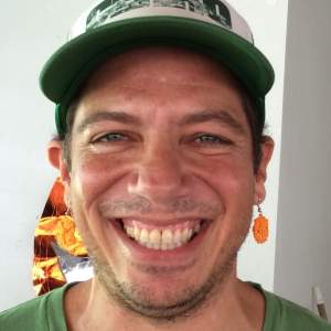 hemi Rosado-Seijo smiles brightly wearing a green shirt, baseball hat, and dangling earring. He is a Puerto Rican man with medium light skin tone, dark hair, and stubble.