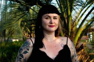 A white woman with light skin tone, black hair, and tattoos stands in front of palm trees, wearing a black top.