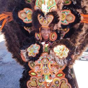 Denise Smith smiles slightly, only her face showing from an elaborate costume with black feathers and colorful beaded designs. She is a Black woman with medium dark skin tone.